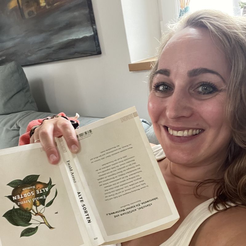 Lisa Sofie Zelgin lacht auf dem Sofa und hält ein aufgeschlagenes Buch in die Kamera