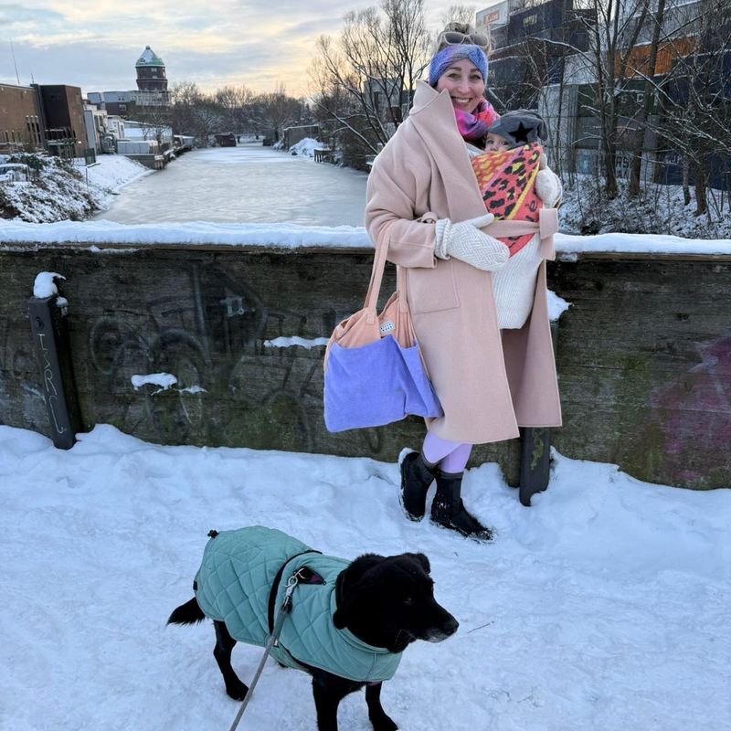 Lisa Sofie Zelgin im Winter auf einer verschneiten Brücke, die Tochter in der Trage, davor ihr schwarzer Hund im grünen Mantel